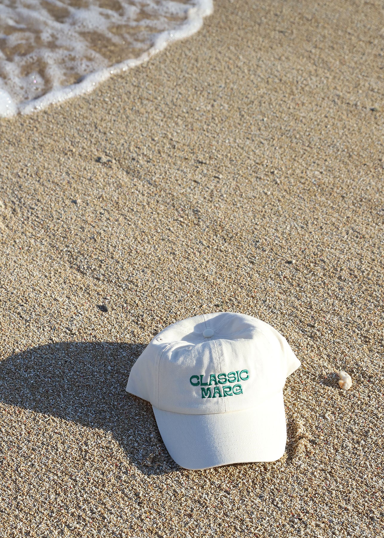 A white 'Classic Marg' dad cap with green embroidery placed on the sand near the water's edge.